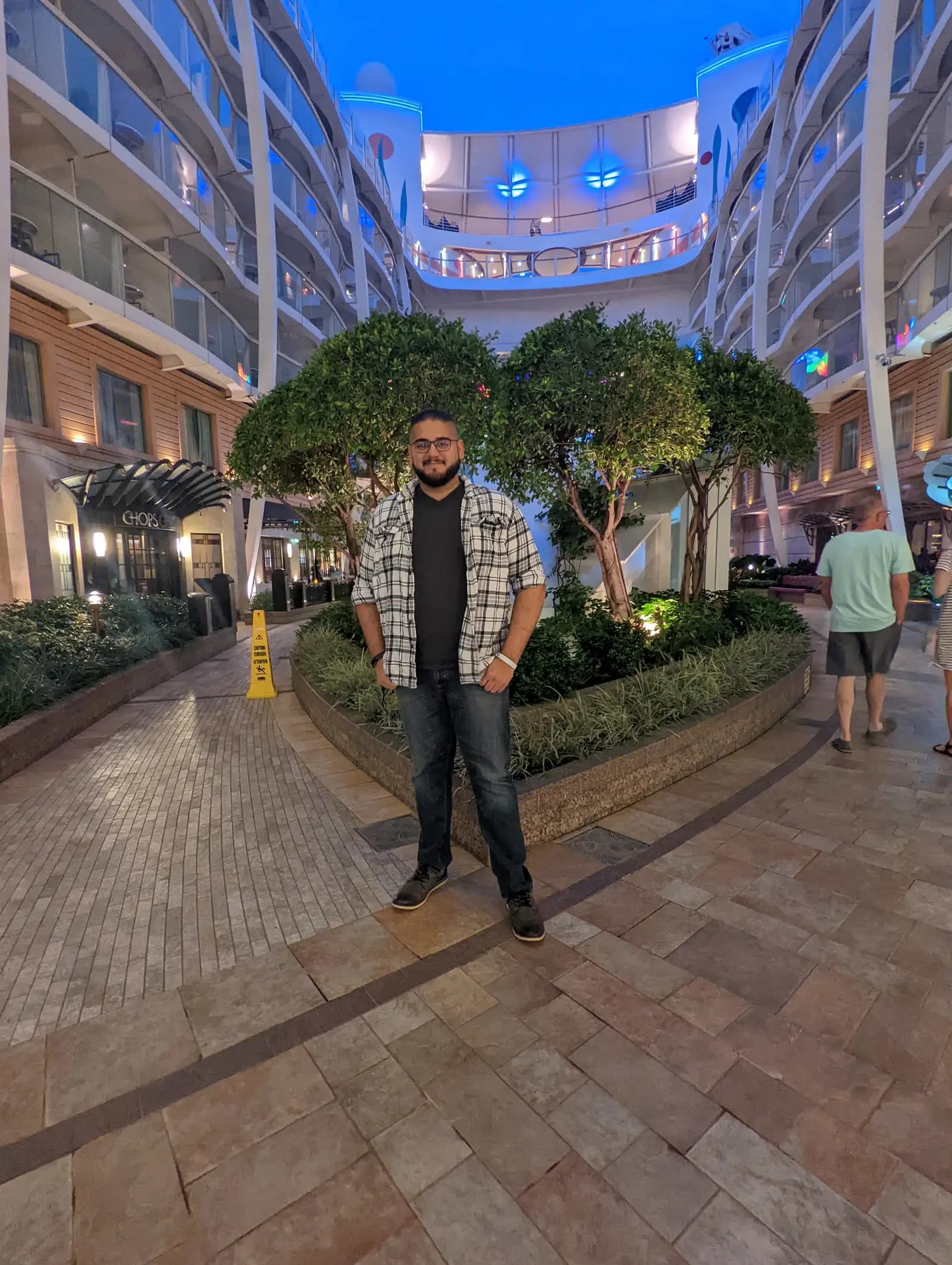 A traveler standing in Central Park on Royal Caribbean's Allure of the Seas at dusk, between lit trees and planters with balcony cabins rising on both sides
