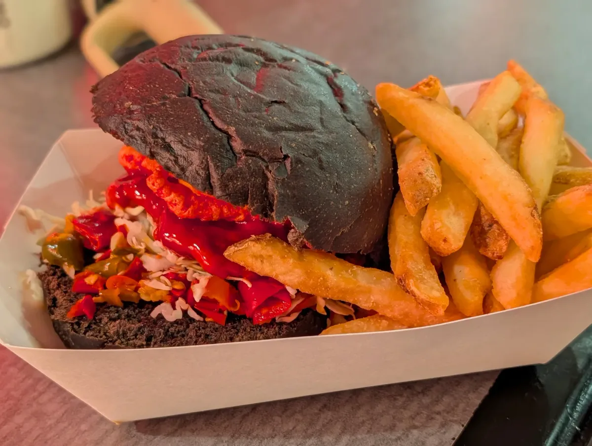 A sandwich on a black bun with red hot sauce, slaw and pickled peppers, served with fries in a paper tray