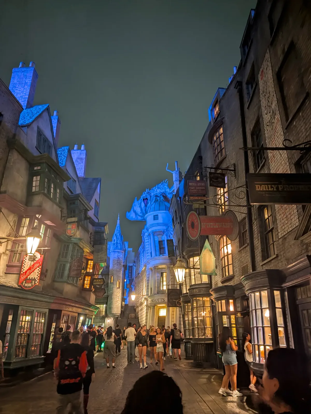 Diagon Alley at night with shop signs lit and the dragon on top of Gringotts glowing blue at the end of the street