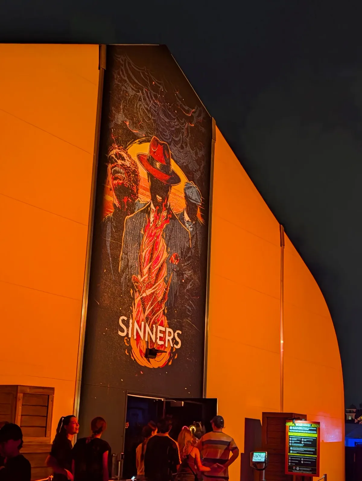 The entrance to the Sinners haunted house at night, with a tall poster of a man in a red fedora and guests waiting at the door